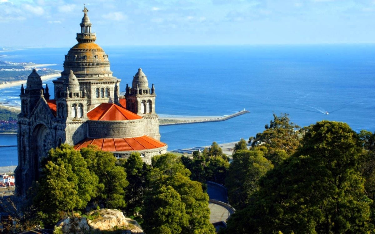 Basílica com cúpula dourada e telhado laranja sobre a paisagem costeira e o mar azul